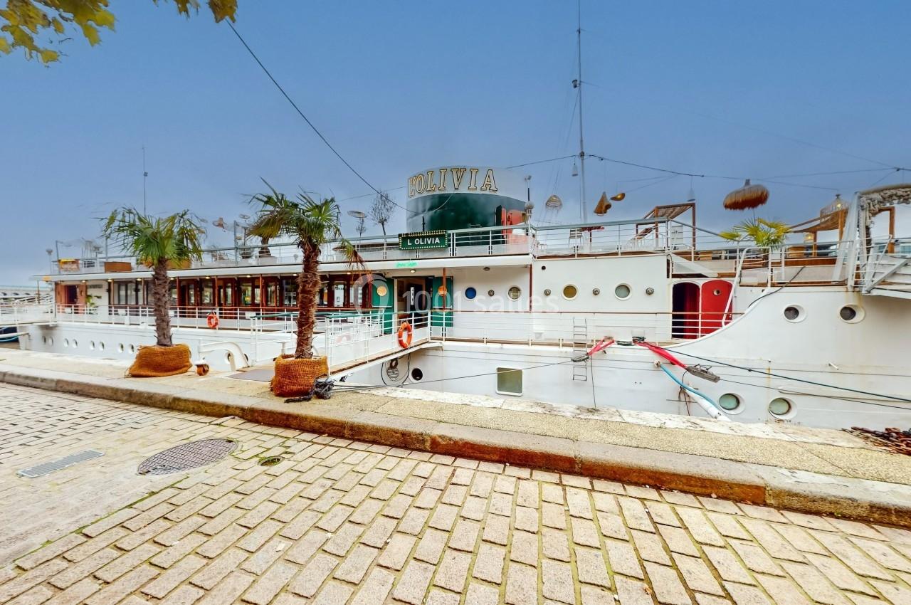 Bateau amarré le long d'un quai pavé, avec des palmiers en pots et un ciel dégagé en arrière-plan.