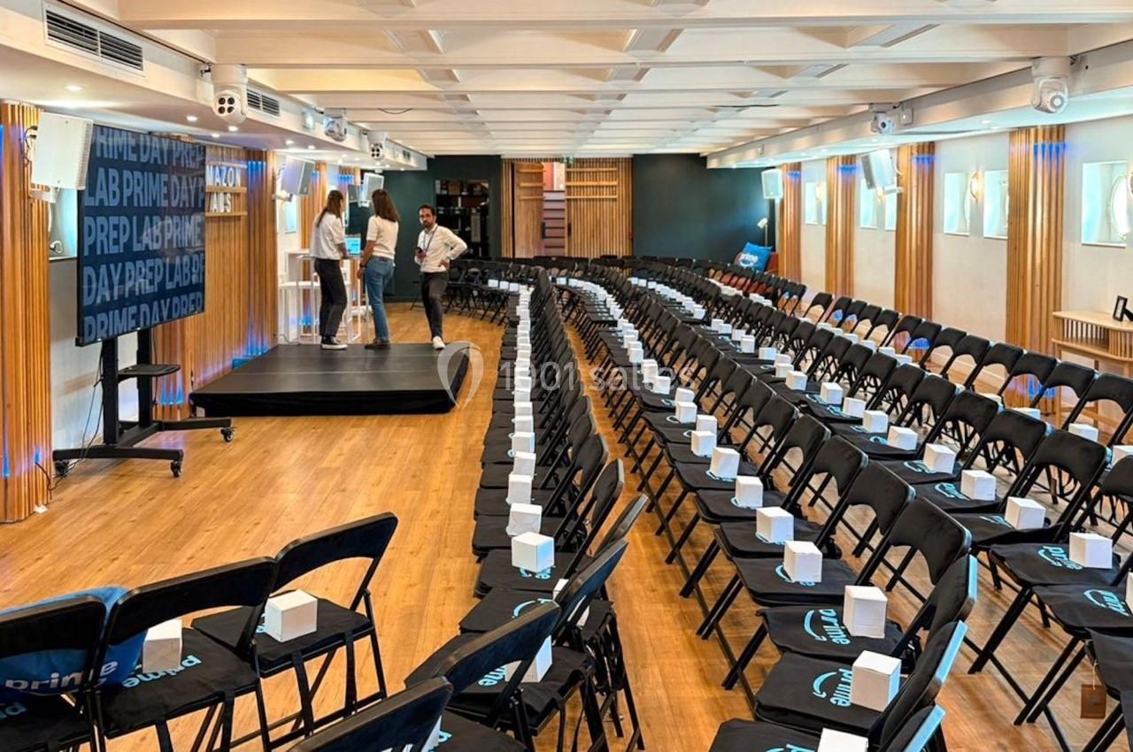 Salle de conférence avec des rangées de chaises noires alignées, une scène et quelques personnes en préparation.
