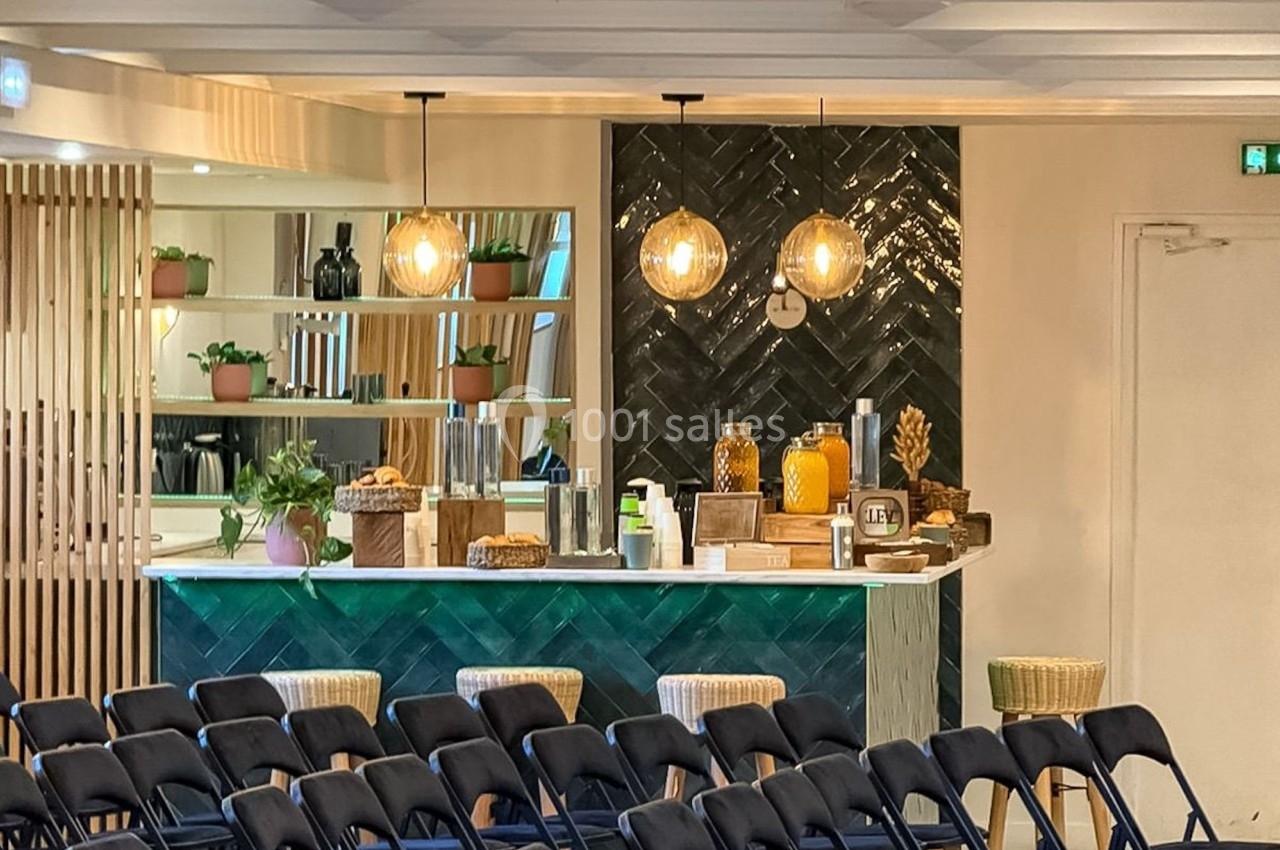 Salle de conférence avec des chaises pliantes alignées face à un comptoir de bar décoré et éclairé par des suspensions.