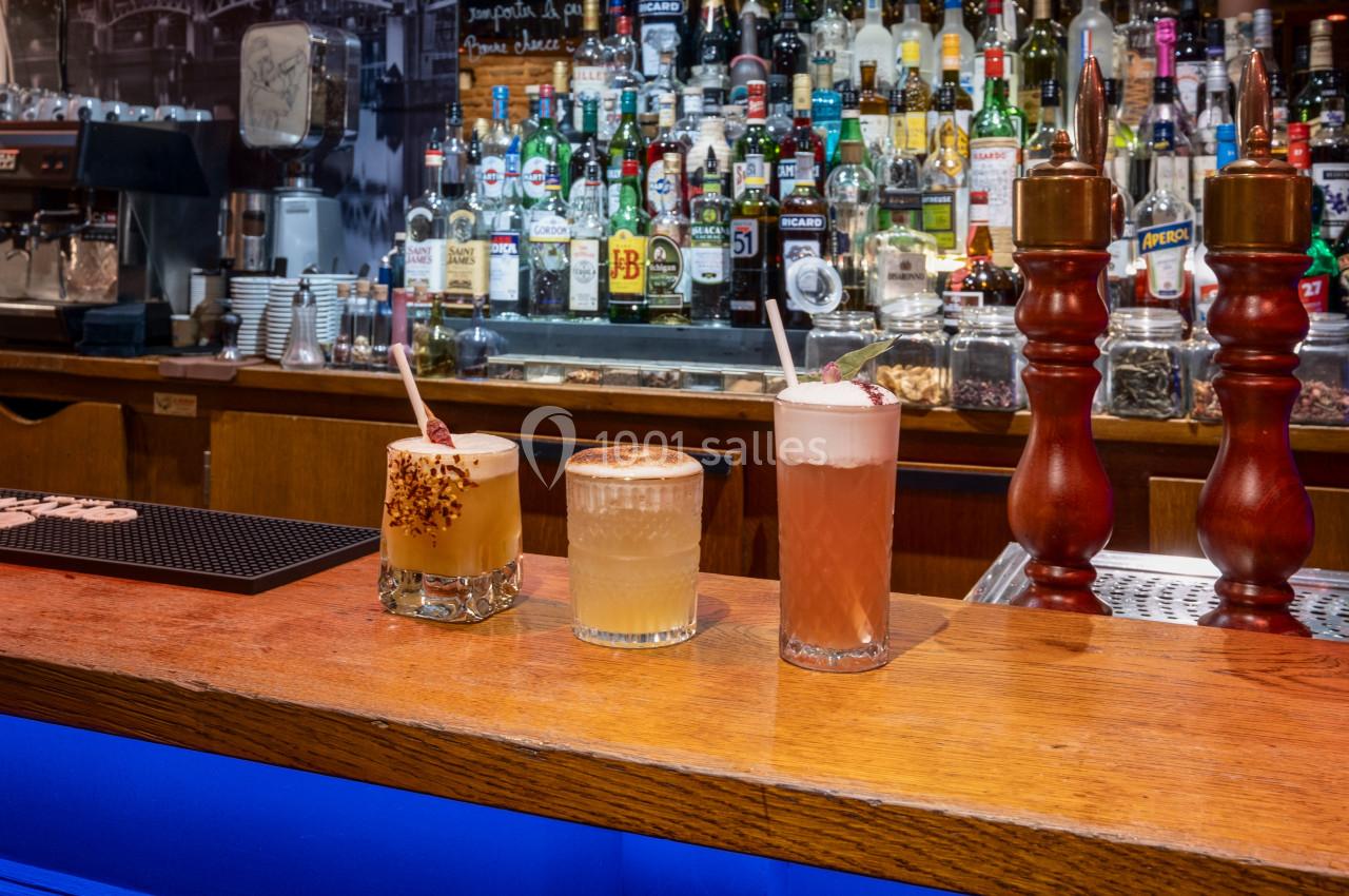 Trois cocktails variés alignés sur un comptoir en bois devant un bar rempli de bouteilles et de tireuses à bière.