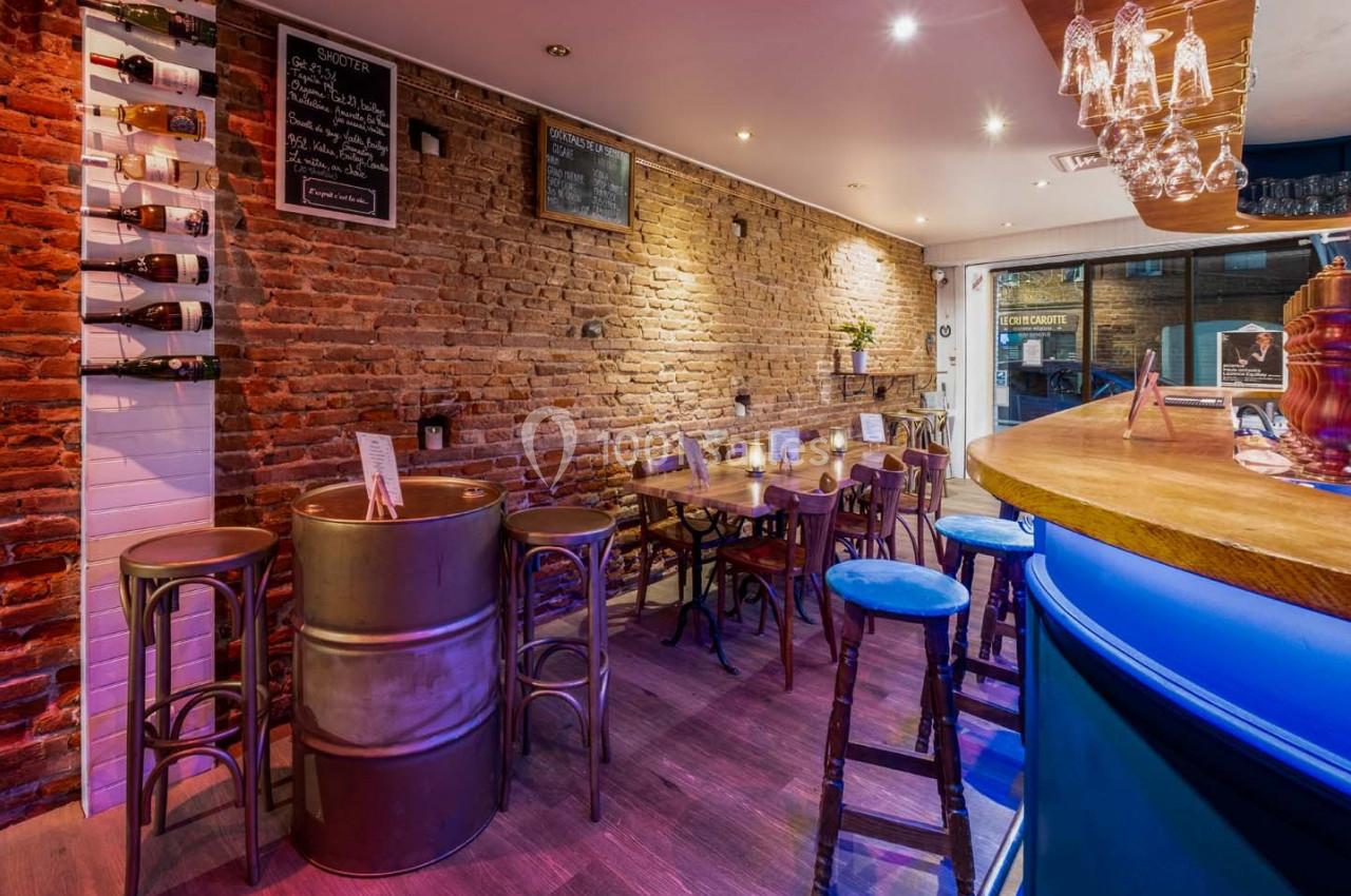 Intérieur d'un bar avec murs en briques, tables en bois, tabourets et comptoir éclairé dans une ambiance chaleureuse.