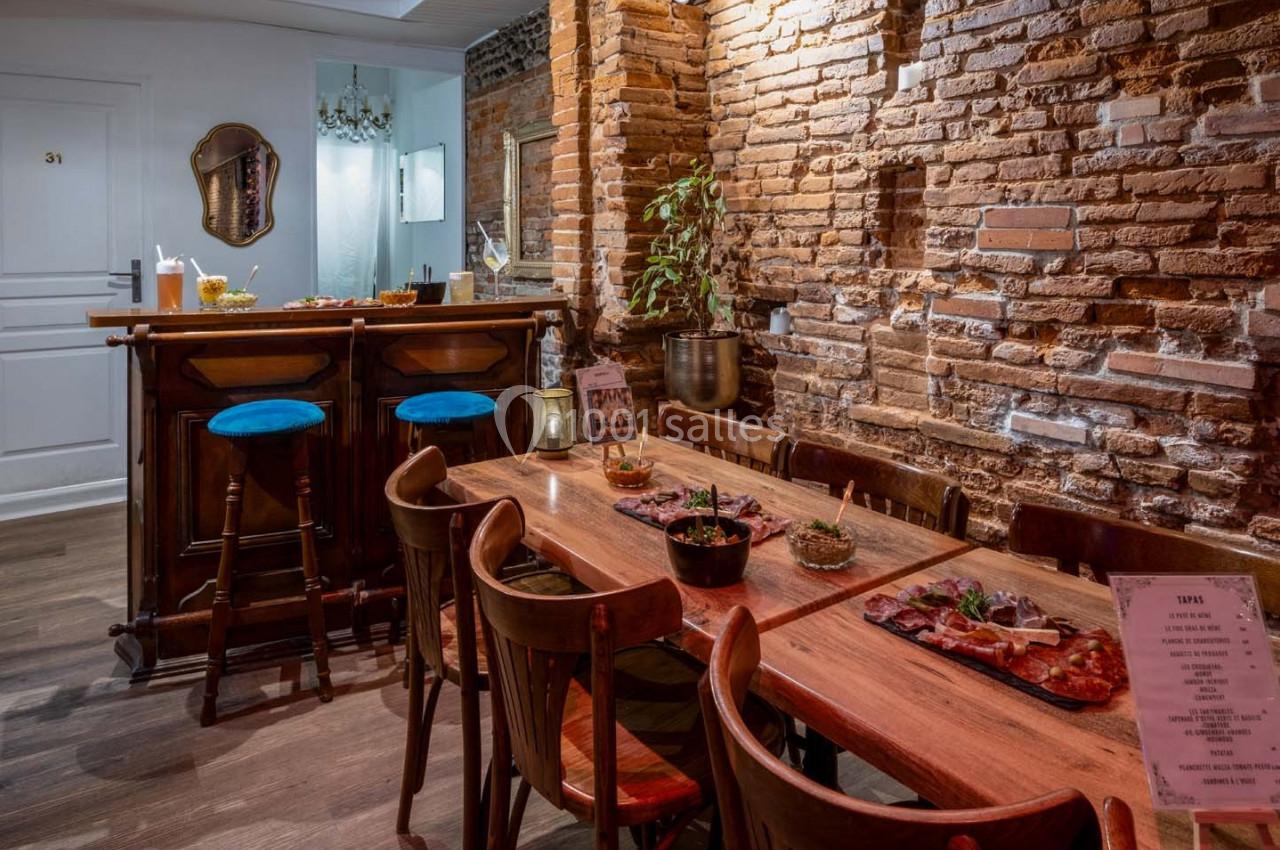 Salle de restaurant avec murs en briques, table en bois dressée, chaises en bois et un bar avec tabourets bleus.