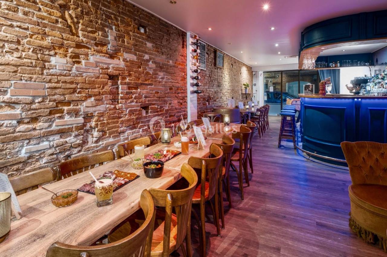 Salle de restaurant avec murs en briques, grande table en bois, chaises alignées et bar éclairé en arrière-plan.