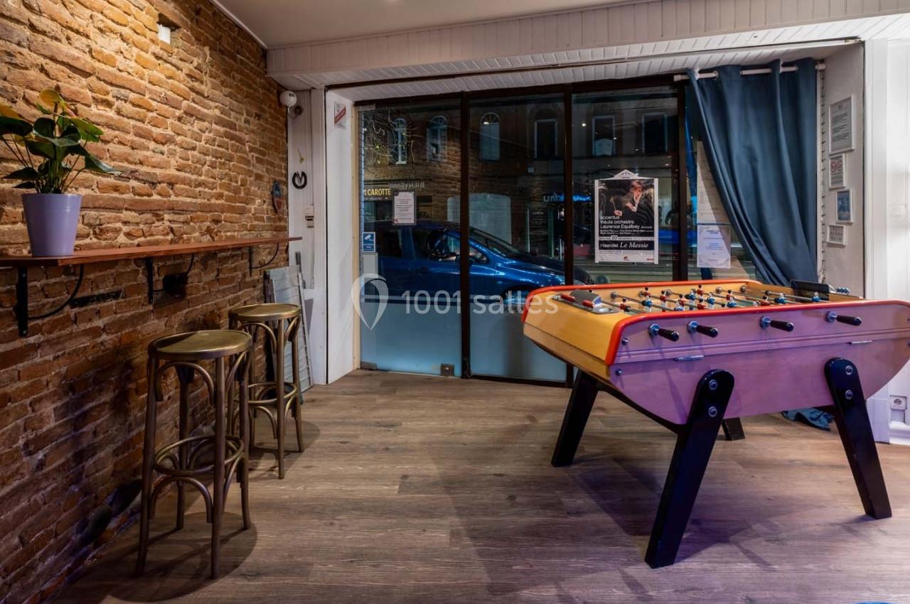 Salle avec un baby-foot, un mur en briques, un comptoir en bois et une grande baie vitrée donnant sur la rue.