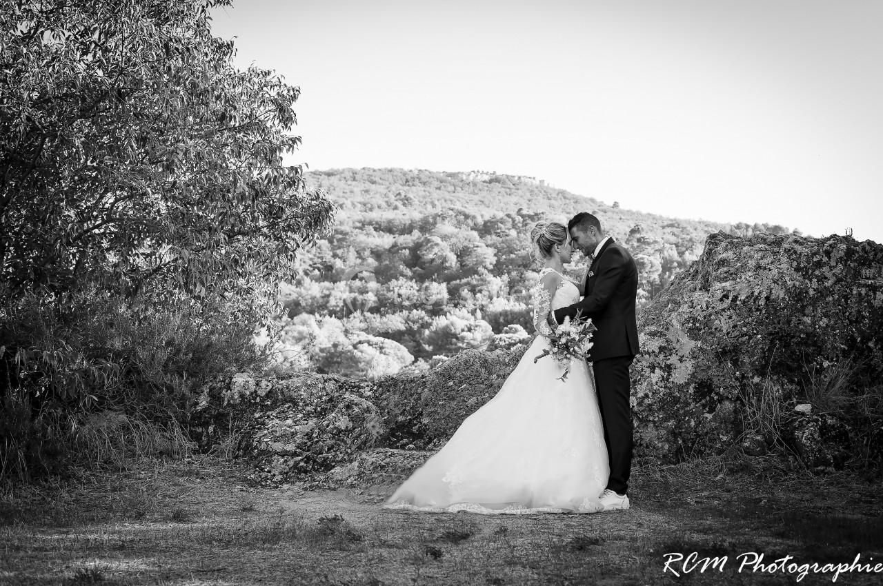 Un couple en tenue de mariage s'embrasse dans un paysage naturel avec des arbres et des collines en arrière-plan.