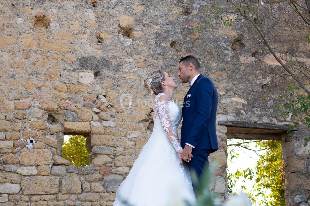 Un couple de mariés se tient la main devant un mur en pierre, avec un bouquet flou au premier plan.
