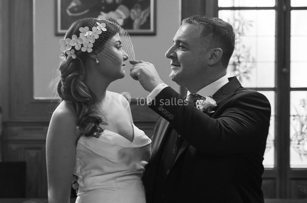Un couple en tenue de mariage sourit dans une salle avec un portrait au mur et une fenêtre en arrière-plan.