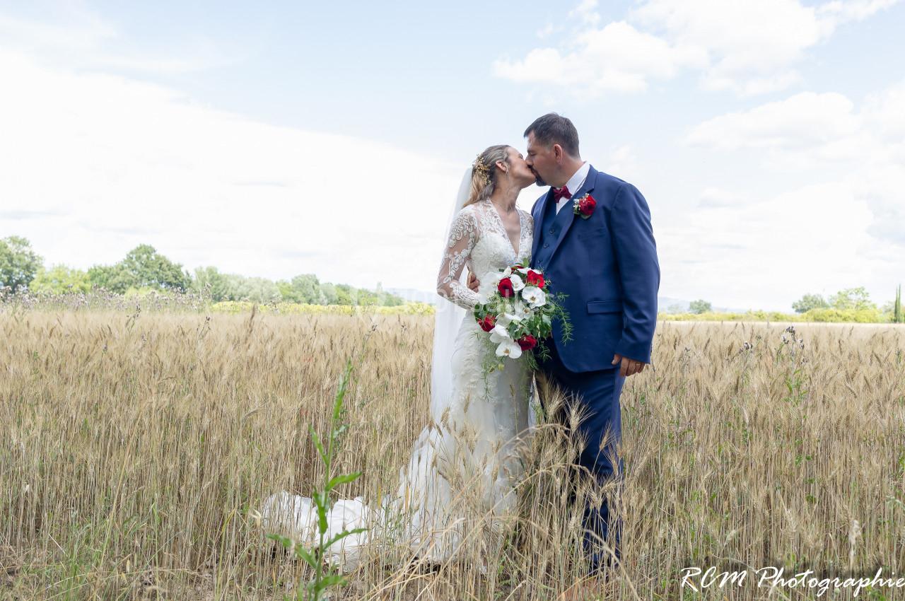 Un couple en tenue de mariage s'embrasse dans un champ de blé sous un ciel partiellement nuageux.