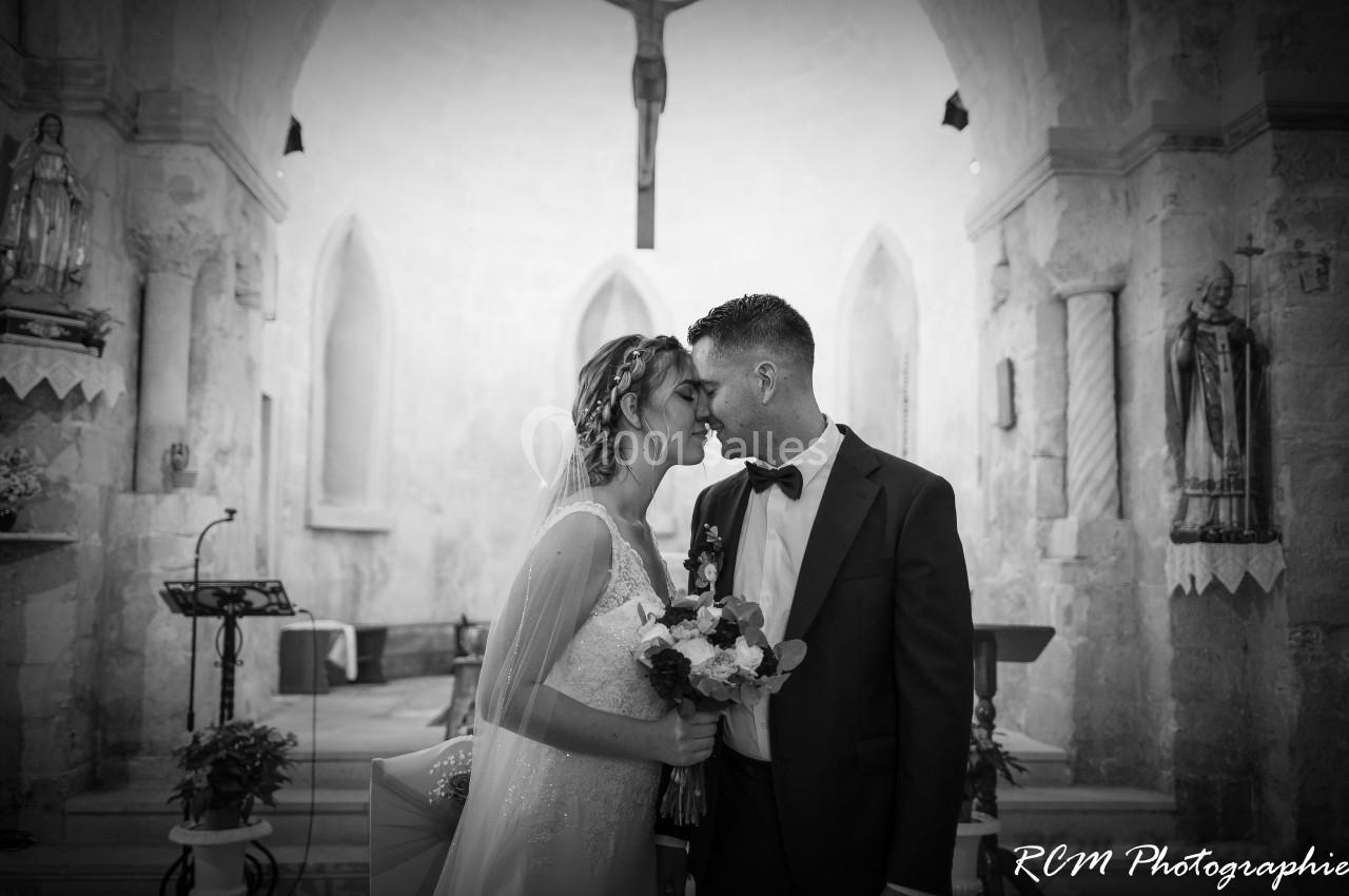 Un couple de mariés se touche le front dans une église, tenant un bouquet, sous un crucifix en arrière-plan.