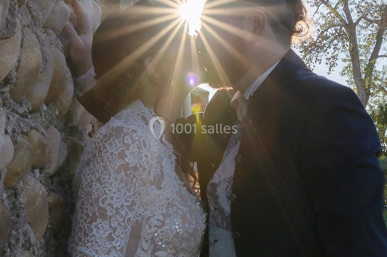 Un couple en tenue de mariage s'embrasse près d'un mur en pierre, éclairé par un rayon de soleil.