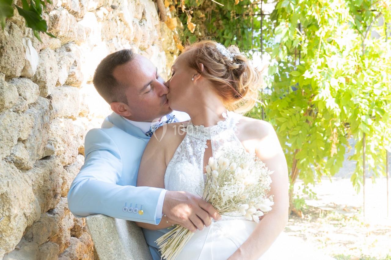Un couple de mariés s'embrasse tendrement près d'un mur en pierre, sous une lumière naturelle.