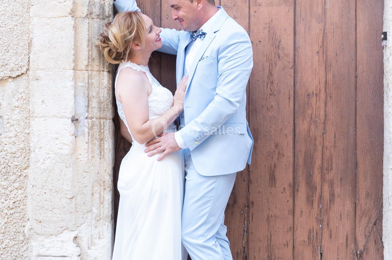 Un couple en tenue de mariage pose devant une porte en bois ancienne, un bouquet posé au sol.