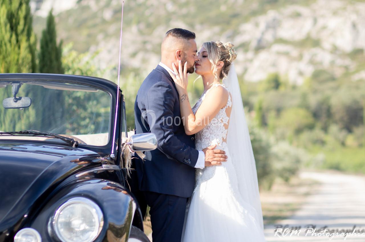 Un couple de mariés s'embrasse tendrement près d'une voiture ancienne dans un paysage naturel.