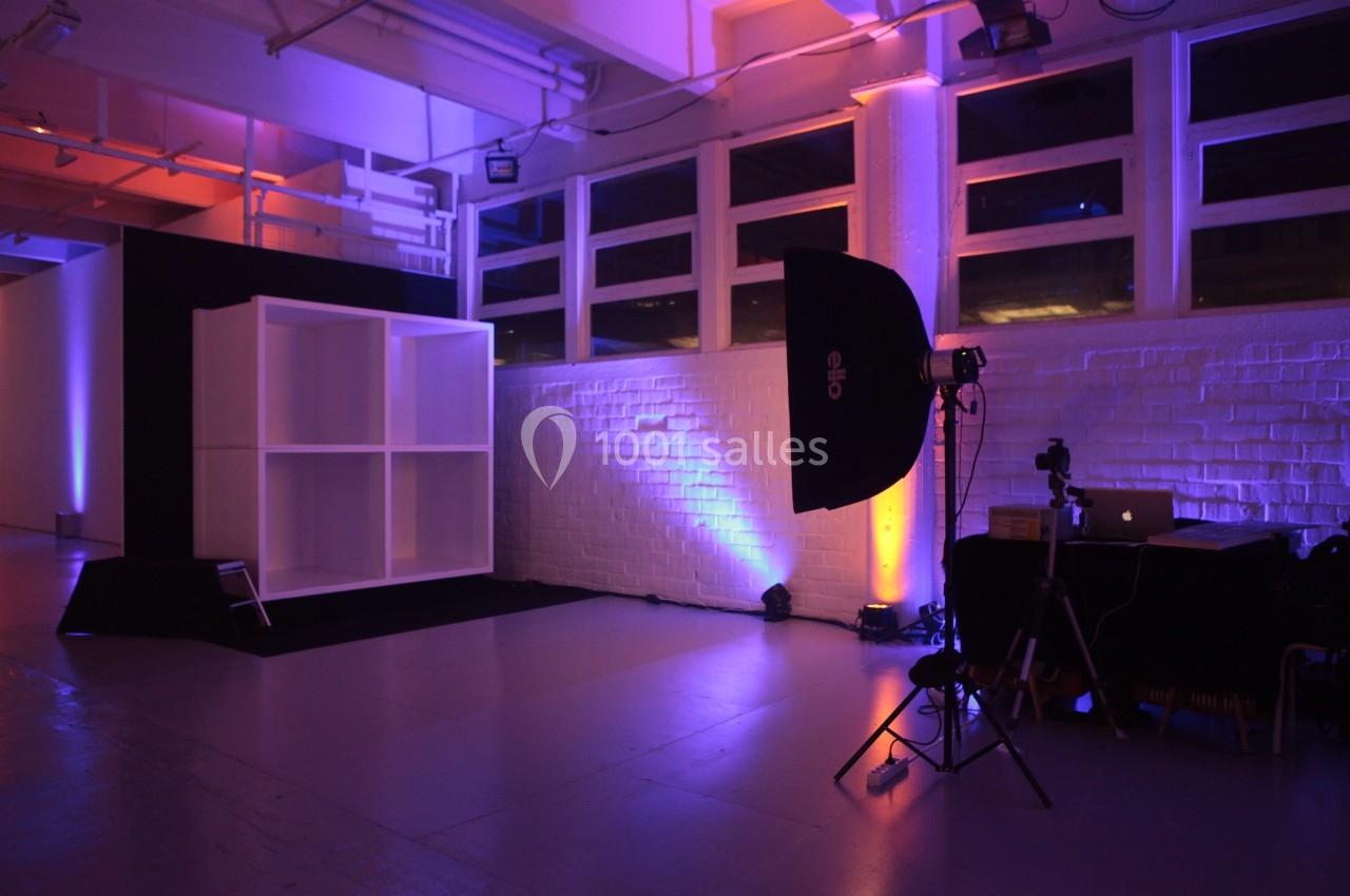 Studio photo avec éclairage coloré, fond blanc, étagère vide, trépied et ordinateur sur une table.