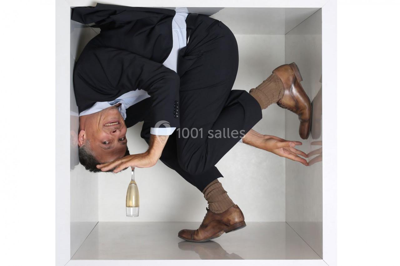 Homme en costume replié dans un espace exigu en forme de cube, tenant une coupe de champagne.