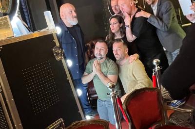 Femme souriante portant un blazer imprimé coloré, un t-shirt gris et un jean, posant devant un fond blanc.