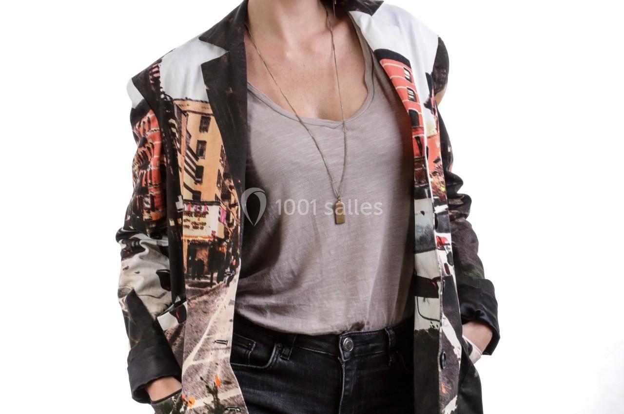 Femme souriante portant un blazer imprimé coloré, un t-shirt gris et un jean, posant devant un fond blanc.