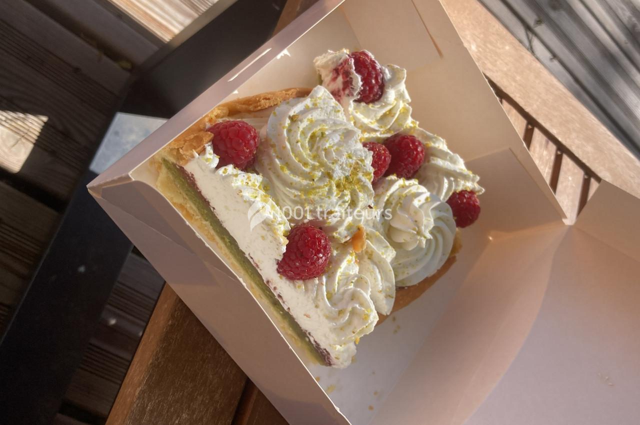 Part de tarte garnie de crème chantilly, framboises fraîches et éclats de pistache, présentée dans une boîte en carton.