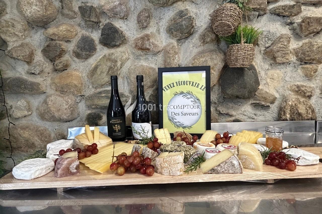 Plateau de fromages variés avec raisins, miel, deux bouteilles de vin et une affiche sur fond de mur en pierre.