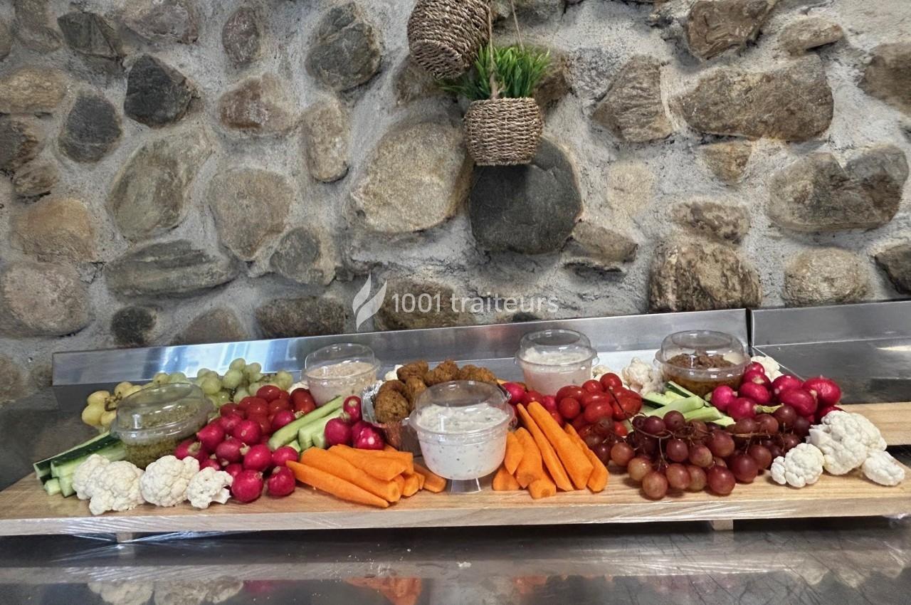 Plateau de légumes frais, fruits et sauces disposés sur une table devant un mur en pierre.
