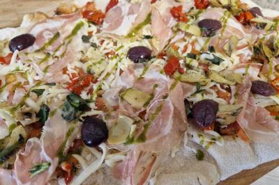 Pizza garnie de jambon cru, olives noires, artichauts, tomates séchées, fromage râpé et filet de pesto.