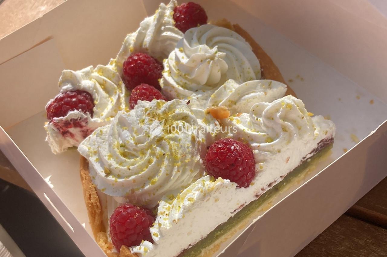 Part de tarte garnie de crème chantilly, framboises fraîches et éclats de pistache dans une boîte en carton.
