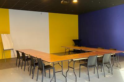 Salle de réunion avec tables en bois disposées en U, chaises noires, mur jaune et tableau de conférence.