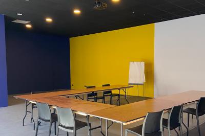 Salle de réunion avec tables en bois disposées en U, chaises noires, mur jaune et tableau de conférence.