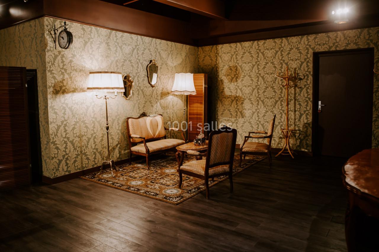 Salon vintage avec papier peint à motifs, mobilier ancien, lampes sur pied et tapis décoratif sur un sol en bois sombre.