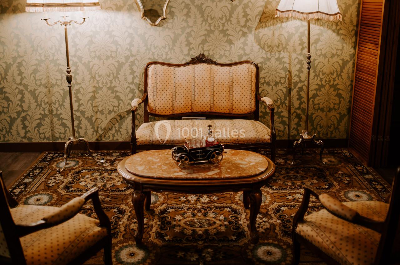 Salon vintage avec canapé, fauteuils, table basse en bois, tapis oriental, lampes et miroirs sur un mur à motifs.