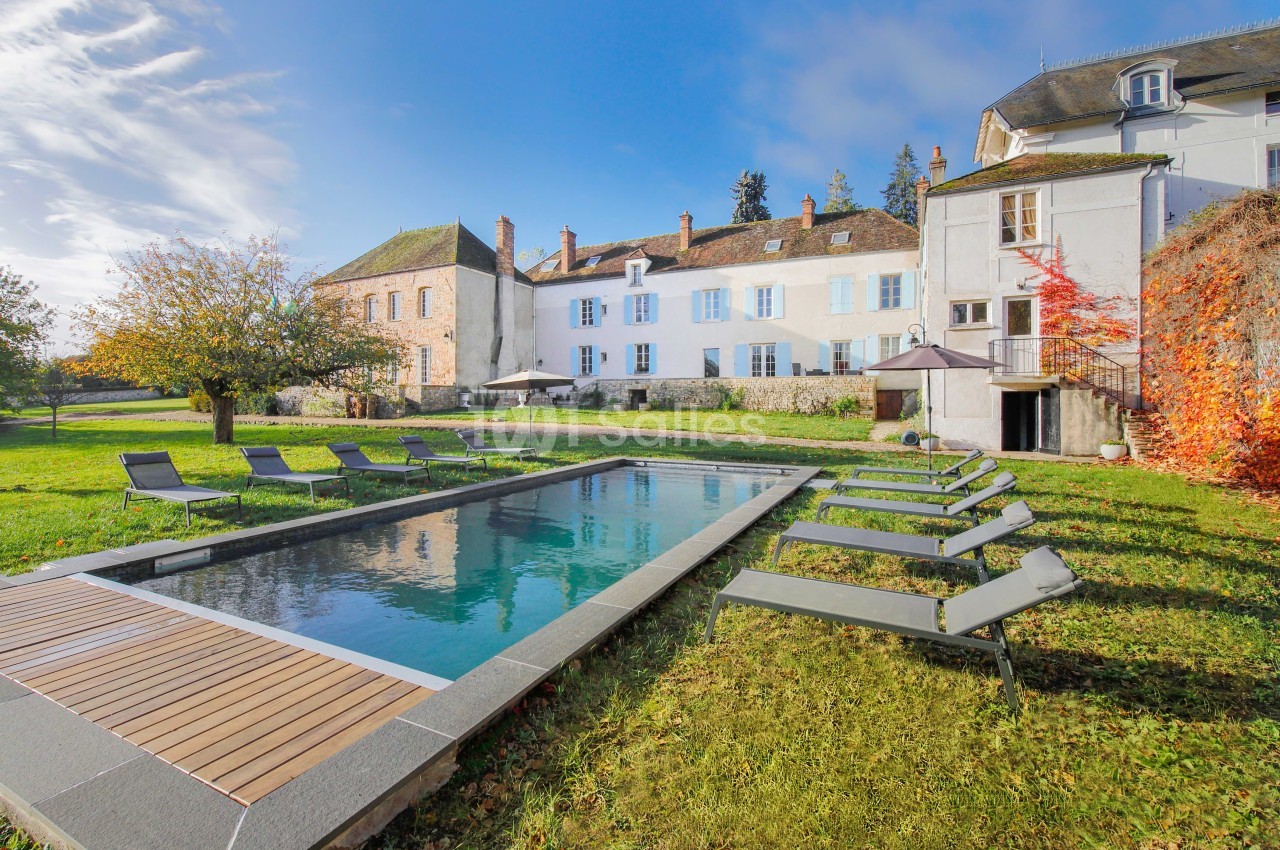 Piscine extérieure entourée de chaises longues dans le jardin d'une grande maison ancienne sous un ciel dégagé.