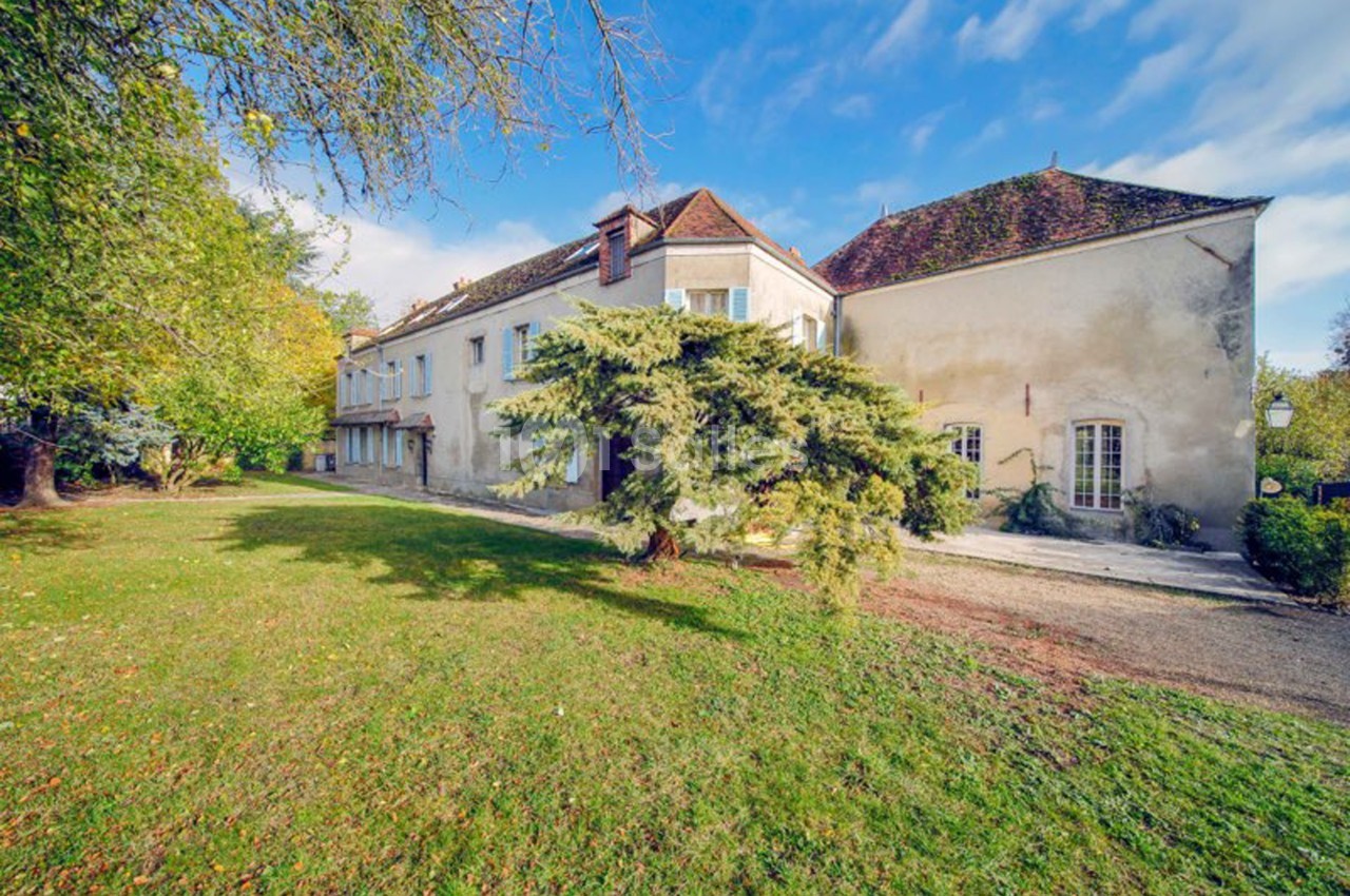 Grande maison ancienne avec toit en tuiles, entourée d'un jardin arboré sous un ciel partiellement nuageux.