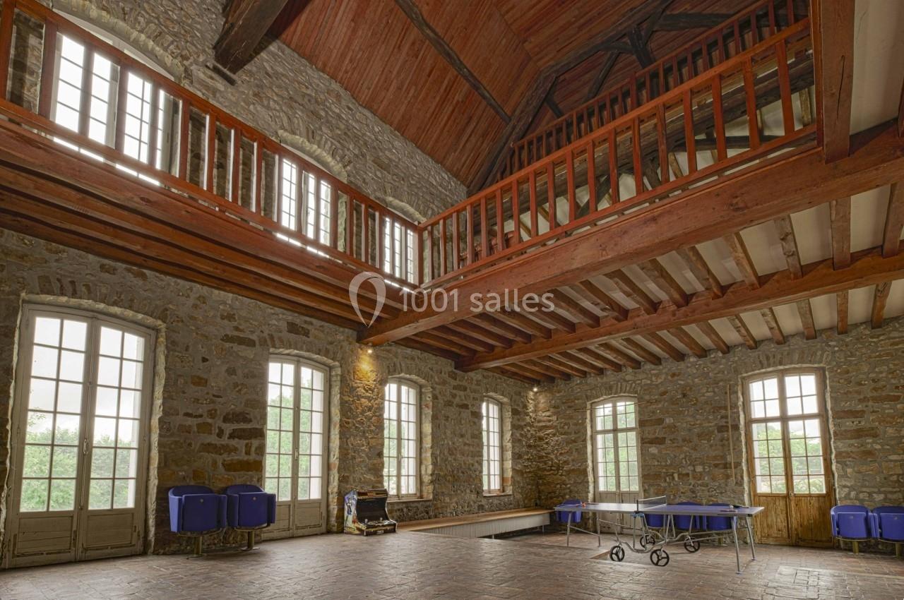Grande salle en pierre avec plafond en bois, mezzanine, grandes fenêtres et tables pliantes rangées au sol.