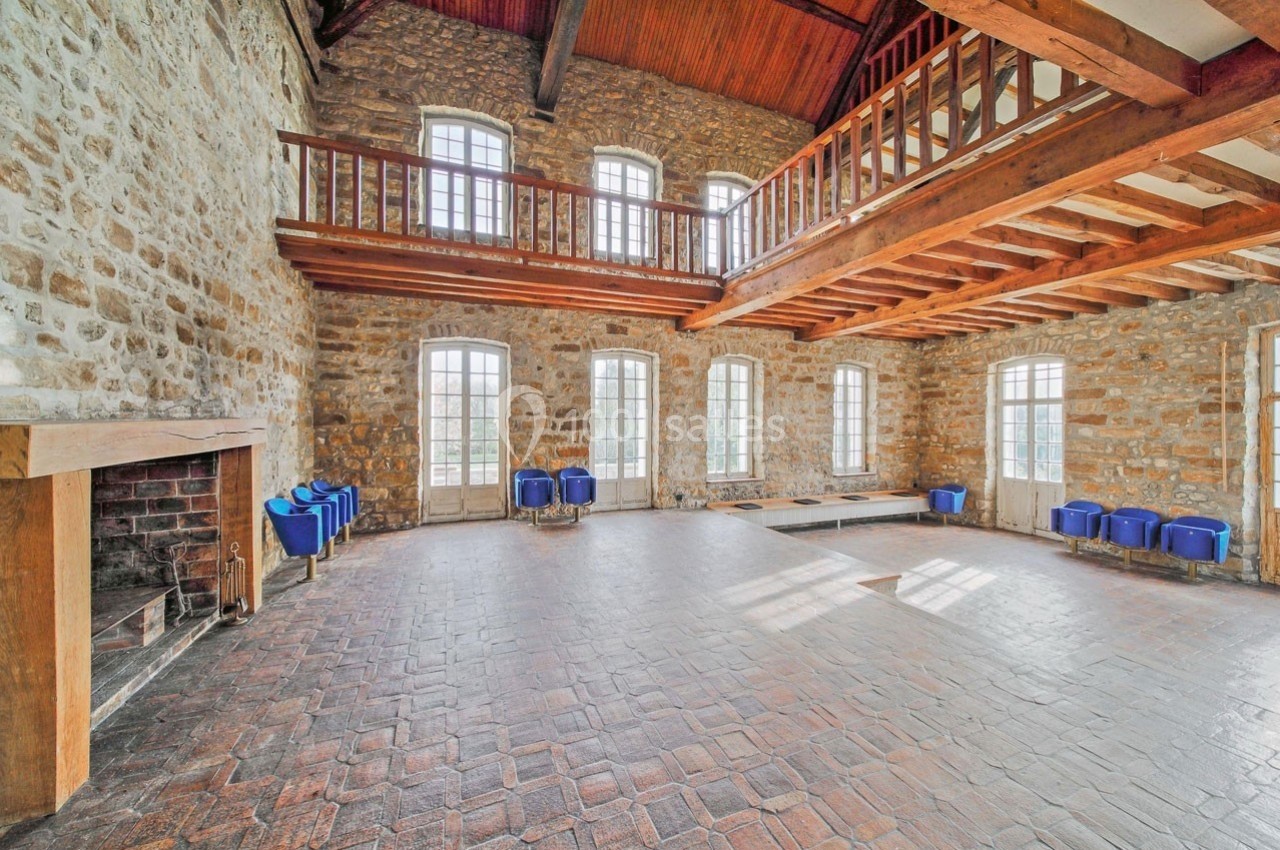 Salle spacieuse avec murs en pierre, plafond en bois, mezzanine et grandes fenêtres laissant entrer la lumière naturelle.