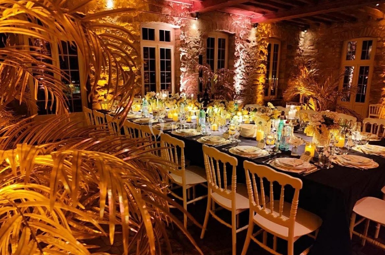 Salle de réception chaleureusement éclairée avec une grande table dressée, entourée de chaises blanches et de décorations…