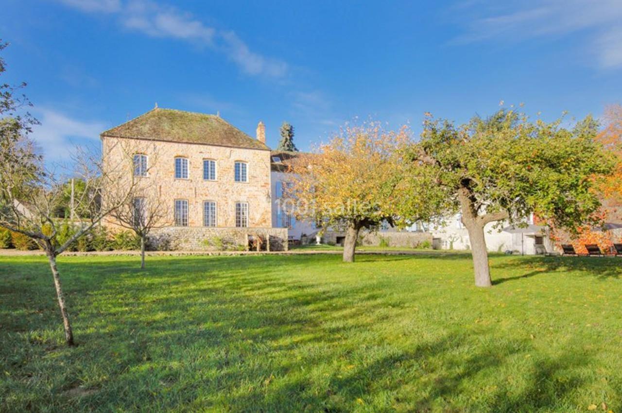 Grand bâtiment en pierre entouré d'un jardin verdoyant avec des arbres, sous un ciel bleu clair.