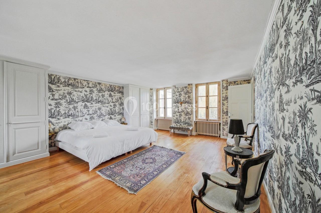 Chambre lumineuse avec lit double, parquet en bois, murs ornés de papier peint à motifs et deux fenêtres.