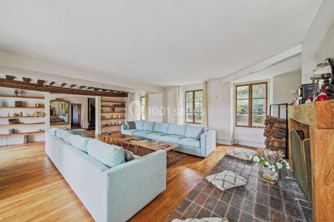 Salon spacieux avec deux canapés bleus, cheminée, parquet en bois et grandes fenêtres laissant entrer la lumière naturelle.