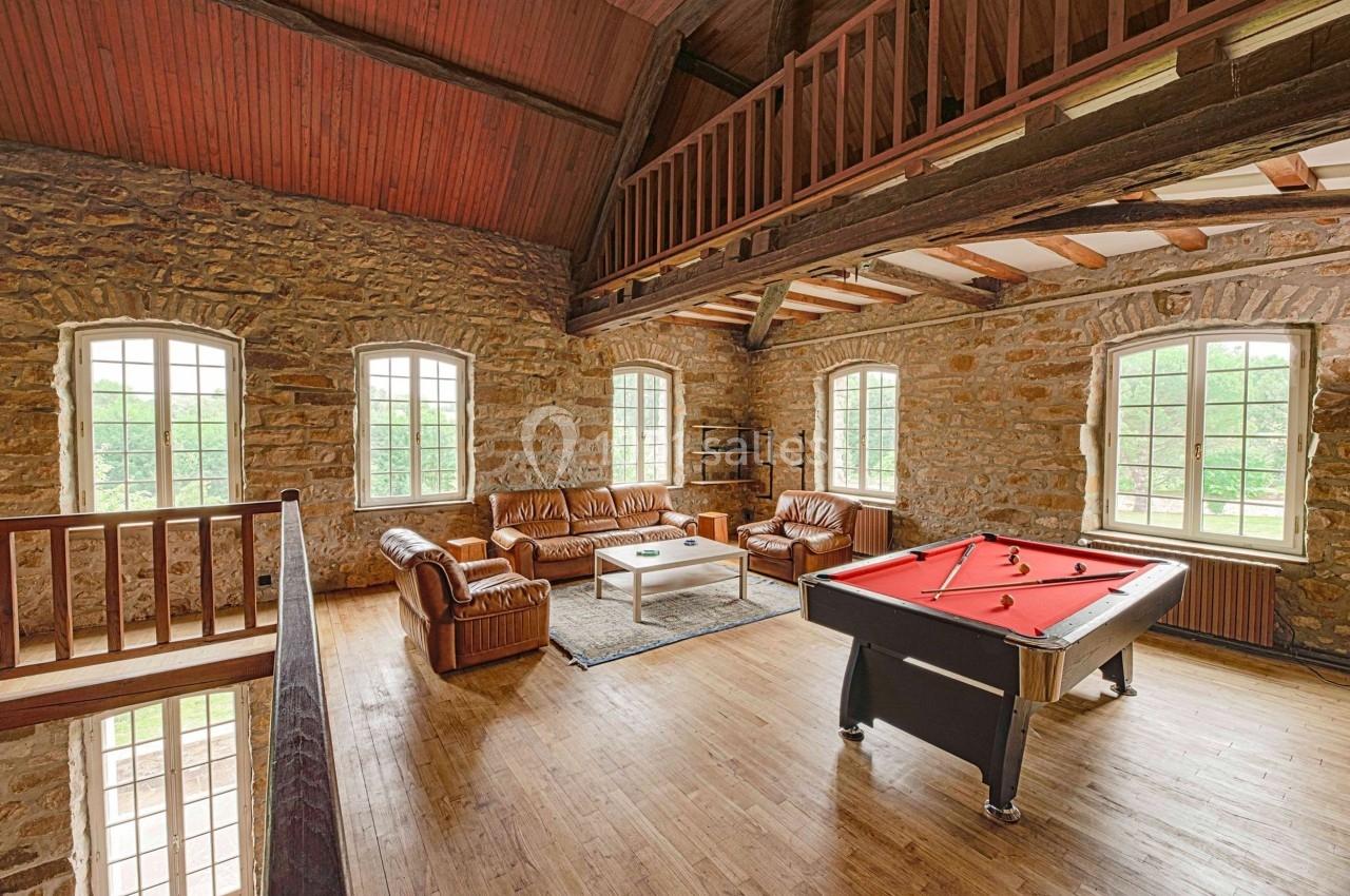 Salle avec murs en pierre, billard rouge, canapés en cuir, table basse et grandes fenêtres laissant entrer la lumière.