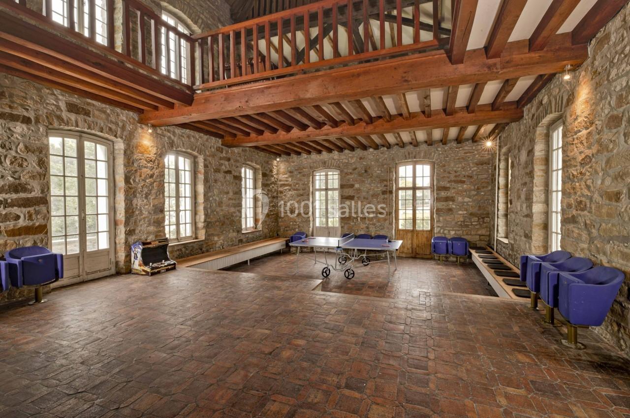 Salle en pierre avec plafond en poutres apparentes, table de ping-pong au centre et fauteuils bleus le long des murs.