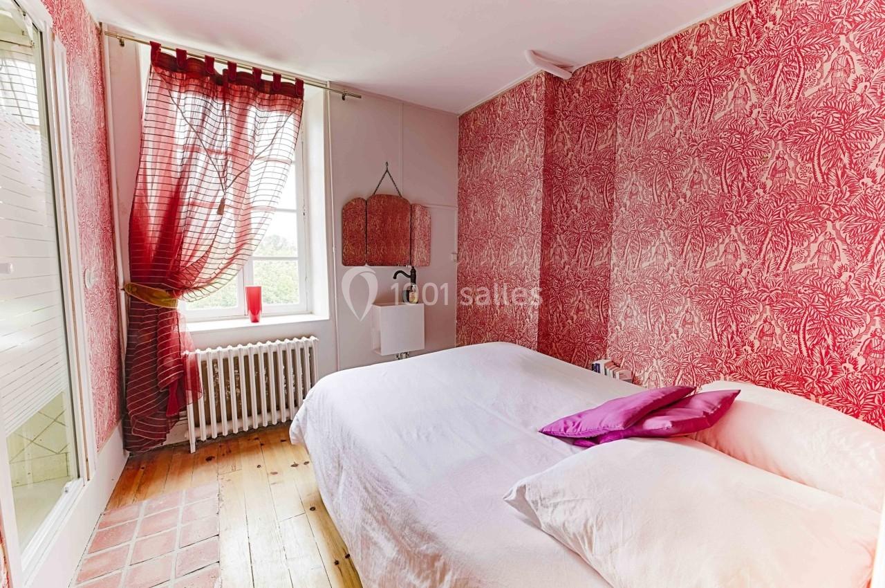 Chambre avec lit double, murs rouges ornés de motifs, parquet en bois et fenêtre avec rideaux laissant passer la lumière.
