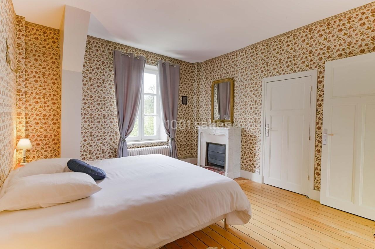 Chambre lumineuse avec lit double, parquet en bois, cheminée décorative, murs à motifs floraux et fenêtre avec rideaux.