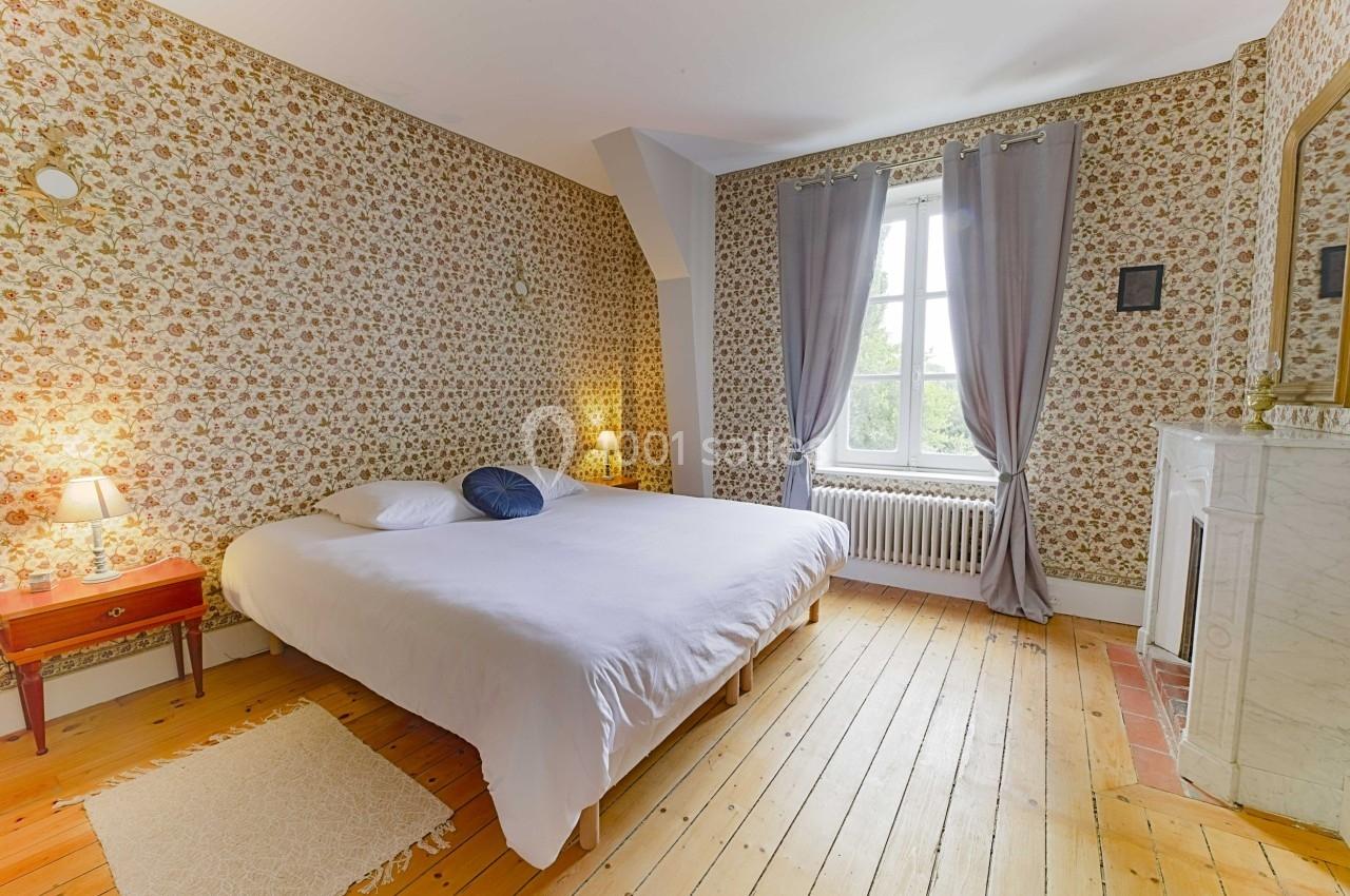 Chambre avec lit double, parquet en bois, papier peint à motifs floraux, cheminée et fenêtre avec rideaux gris.