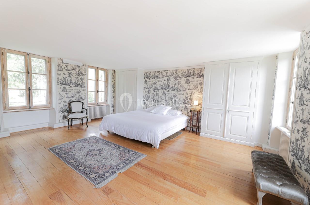 Chambre lumineuse avec lit double, parquet en bois, murs tapissés, fenêtres à carreaux et mobilier classique.