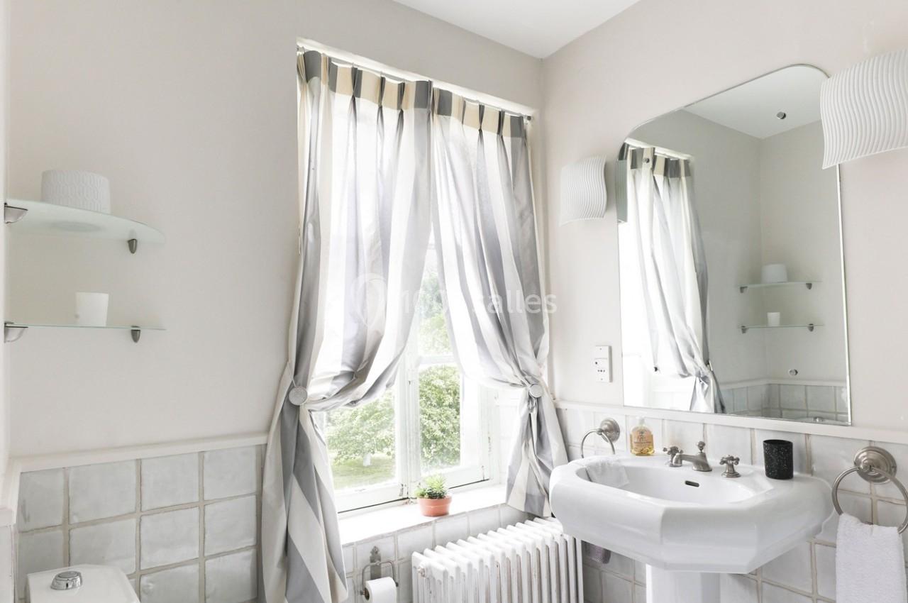 Salle de bain lumineuse avec lavabo, toilettes, miroir, carrelage blanc et fenêtre habillée de rideaux rayés.