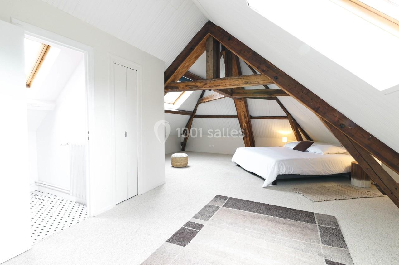 Chambre sous les combles avec poutres apparentes, lit double, tapis et lumière naturelle provenant de fenêtres de toit.