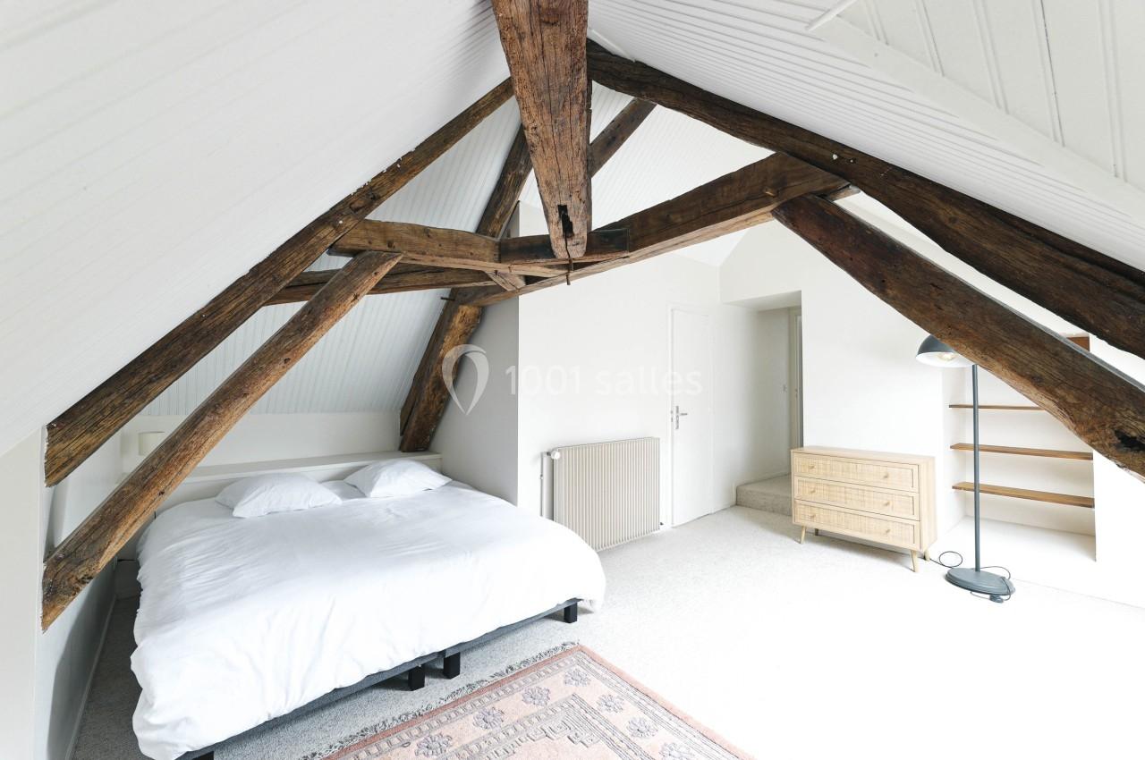 Chambre mansardée lumineuse avec poutres apparentes, lit double, commode en bois, étagères et lampadaire.