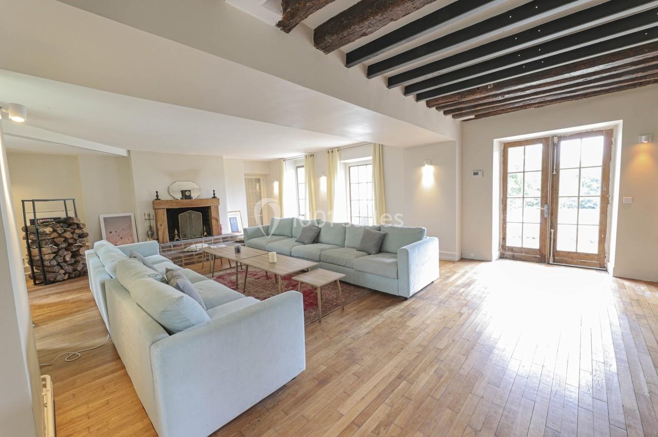 Salon spacieux avec parquet en bois, canapés clairs, cheminée, poutres apparentes et grandes portes-fenêtres.