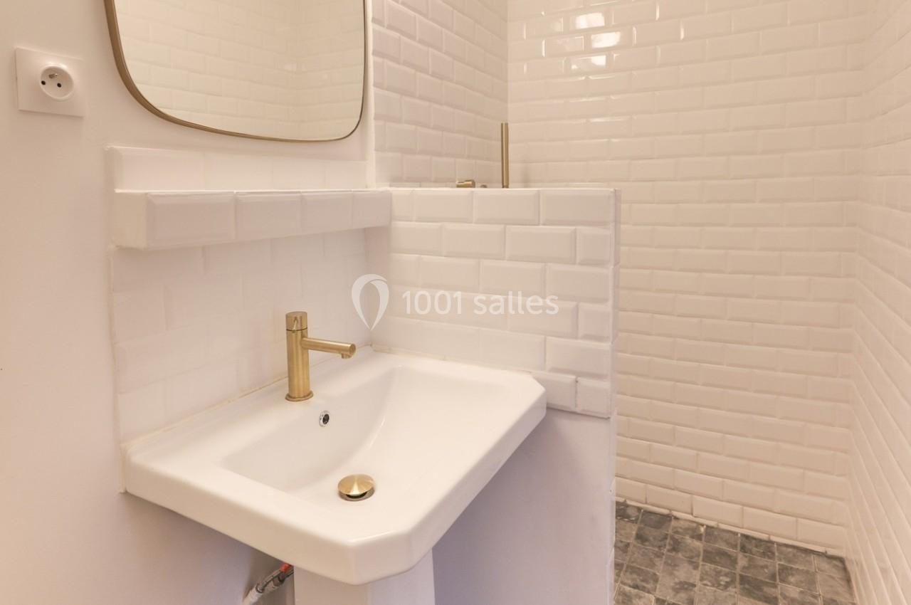 Lavabo blanc avec robinet doré, miroir rectangulaire et murs en carrelage blanc dans une salle de bain minimaliste.