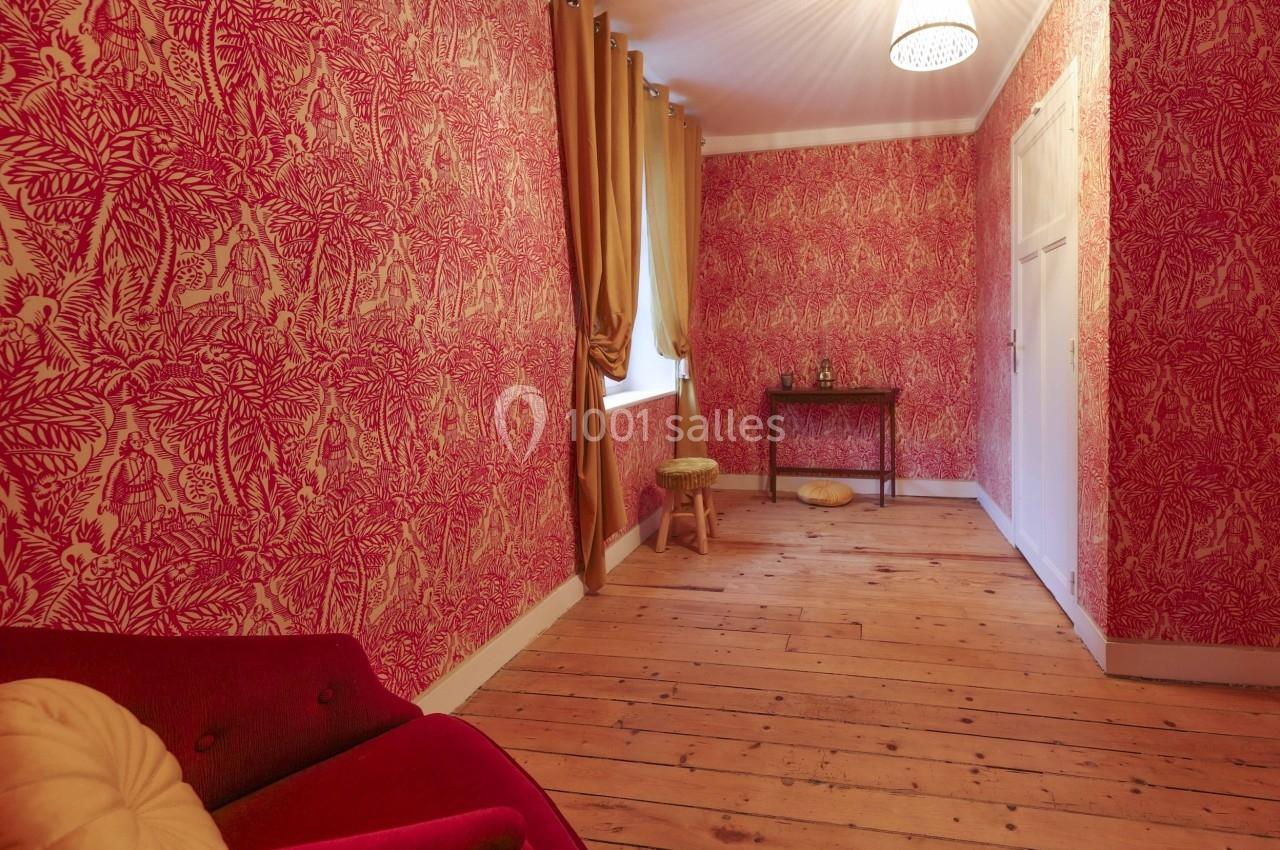 Pièce avec parquet en bois, murs recouverts de papier peint rouge à motifs, rideaux dorés et petit mobilier.